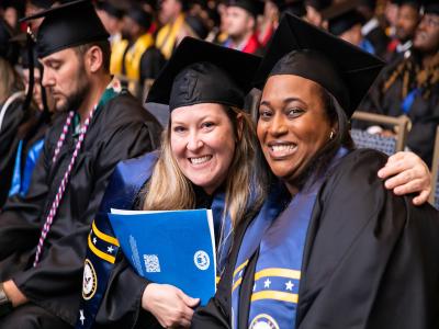 focus on two graduates wearing caps and gowns with other graduates in the background