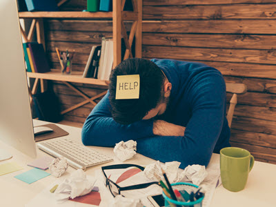 person with their head down on a desk and a sticky note on them that says: help