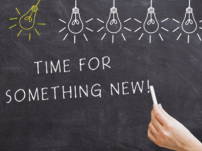 one yellow lightbulb and four white light bulbs drawn in chalk on the top of a chalkboard with the words written by a hand with chalk saying: Time for Something New!