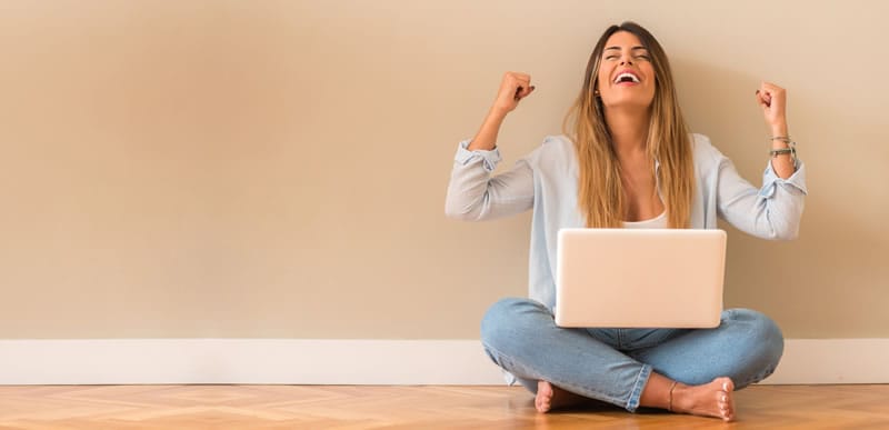 a person sitting on the floor with a laptop and arms up in a celebratory pose