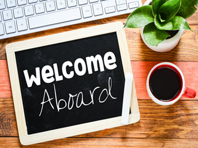 Welcome Aboard chalkboard in front of a keyboard and beside a plant and cup of coffee