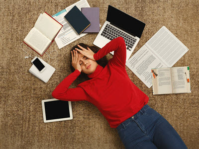 Person who appears to be overwhelmed and lying on the floor surrounded by a tablet, notebooks, books, a phone, and laptop